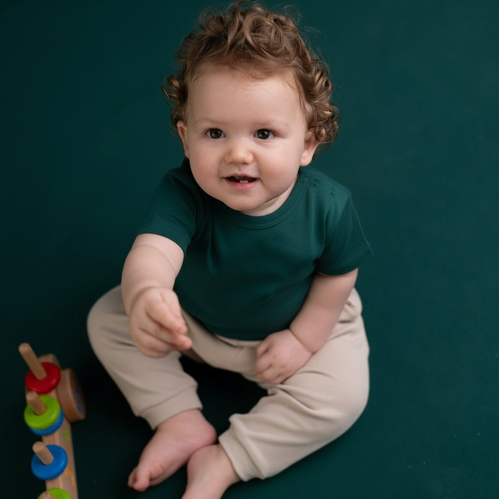 A baby wearing a green organic cotton t-shirt with snap buttons at the neckline, pointing at a colorful wooden toy with a smile on the baby's face.